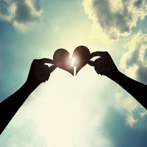 Two hands hold a broken paper heart against a cloudy sky with sunlight shining through.
