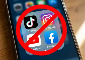 A close-up photograph of a smartphone screen displaying the app icons for TikTok, Instagram, YouTube, and Facebook, with a large red circle and diagonal line ban symbol superimposed across all four icons.