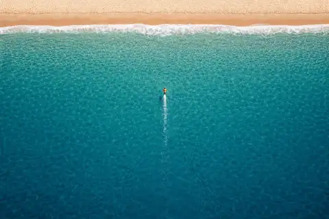 Landscape aerial drone view of a tiny lone swimmer in the open ocean heading straight toward a sandy beach at the top of the frame, leaving a narrow wake behind across a vast expanse of blue water.