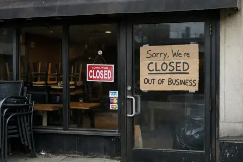 A street-level photo of a recently closed restaurant with dark windows, stacked chairs inside, a closed sign, and a handmade cardboard “out of business” notice taped to the glass door.