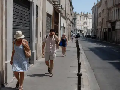 A quiet French city street during a severe heat wave, with closed shop shutters, pale buildings, hard sunlight, and a few people walking slowly in summer clothes.