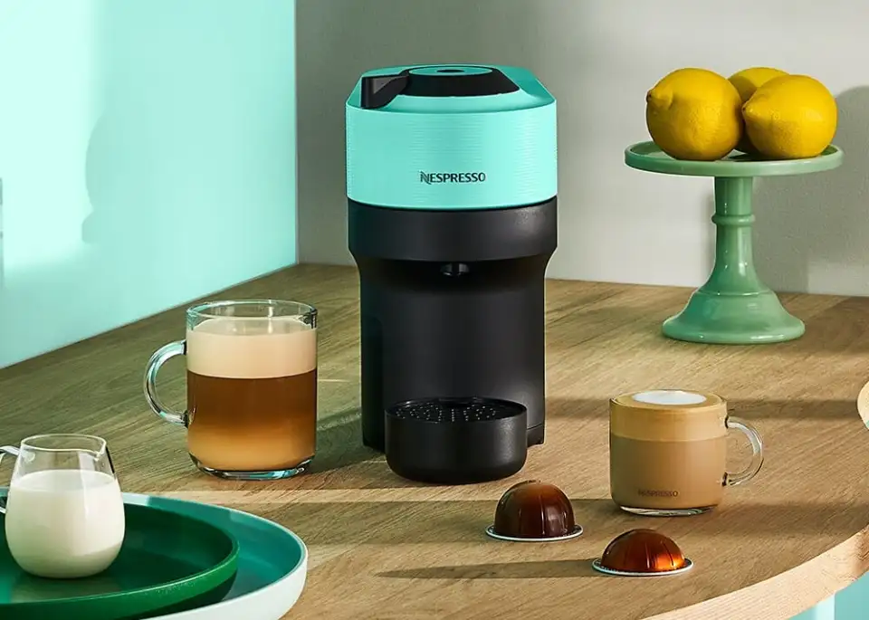 A black and gray Nespresso Coffee machine on a white background. There is a glass of coffee in front of the machine, and to the right are several unused coffee pods.